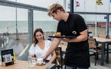 Jonah Kamstra serveert Achsa de Bruijn uit Tholen een koffie met appelgebak op het terras van restaurant Land- en Zeezicht op de pier bij Holwerd.