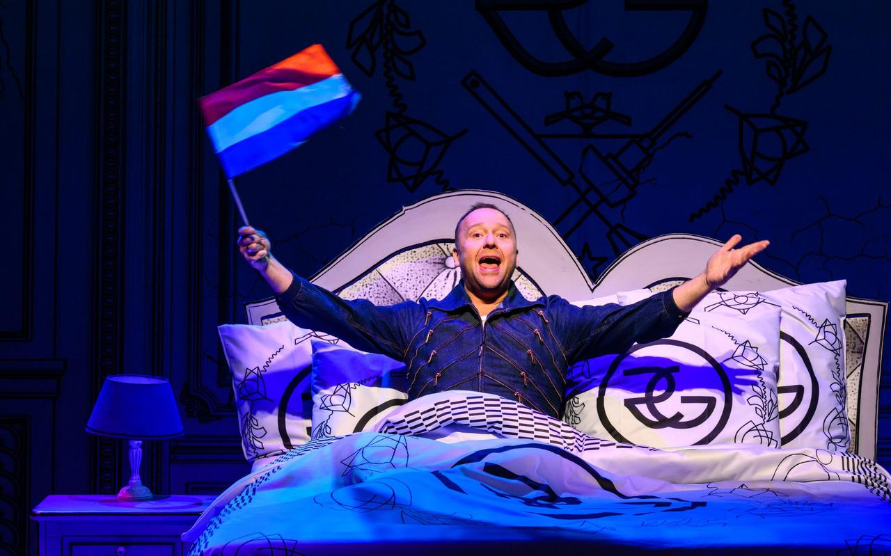 Richard Groenendijk opent zijn show liggend in een kingsize bed, in vol koninklijk ornaat met een kroonluchter boven hem en een majestueus logo op sprei, kussens en hoofdeinde.